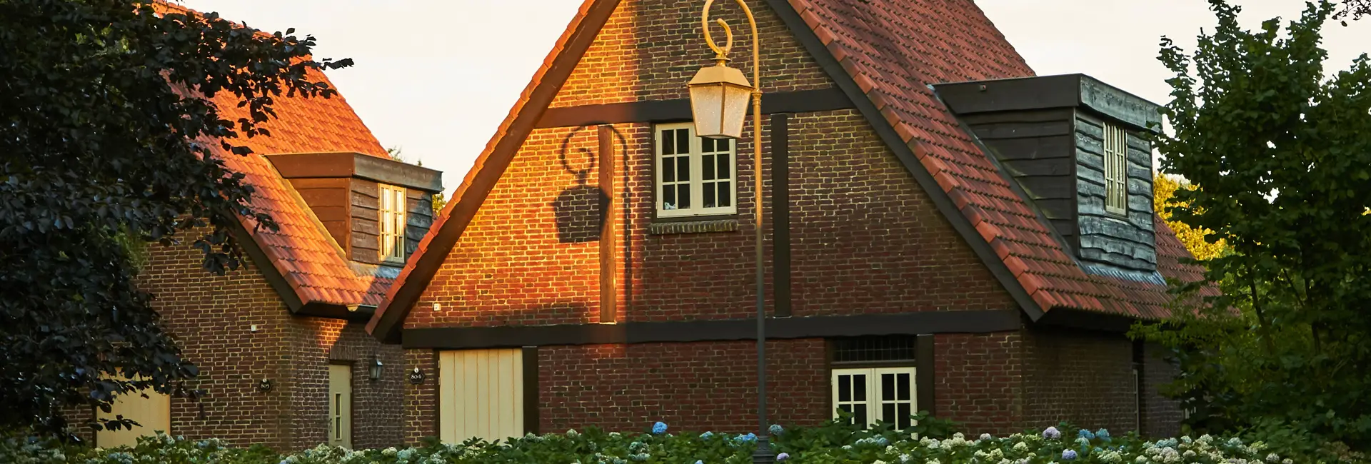 Ein Backsteinhaus mit steilem roten Dach, beleuchtet vom Sonnenlicht. Ein Laternenpfahl wirft einen Schatten an die Wand. Im Vordergrund stehen Büsche mit blauen Blüten.