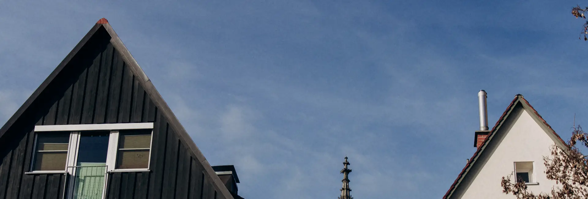 Zwei Hausdächer vor einem blauen Himmel, eines mit einer steilen schwarzen Fassade und ein anderes mit einer weißen Wand und einem Schornstein, der teilweise von einer Turmspitze verdeckt wird.
