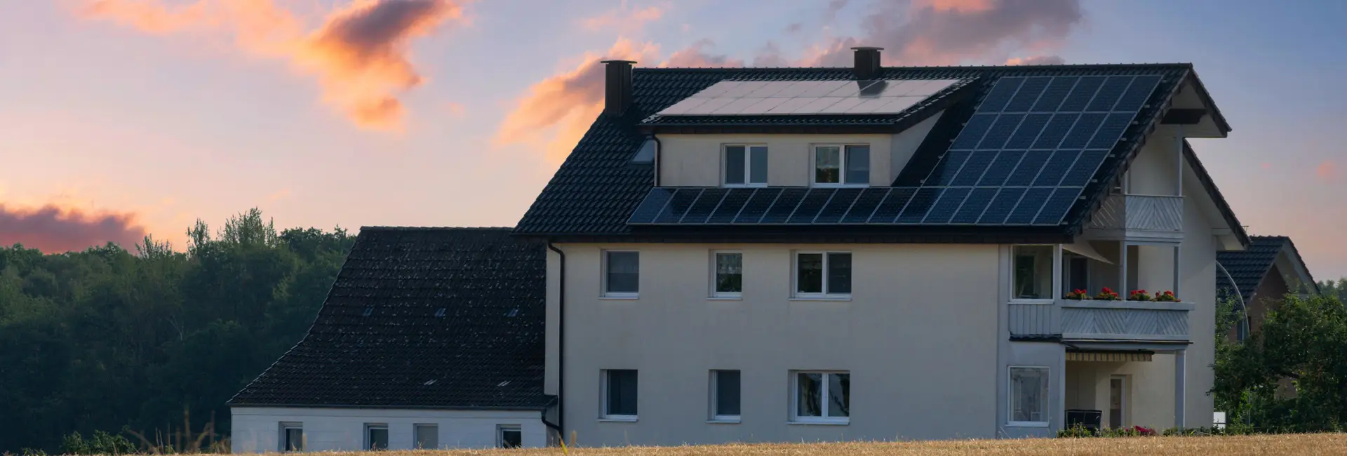 Ein Haus mit Solarmodulen auf dem Dach steht in einer ländlichen Gegend. Der Himmel ist rosa und orange, mit ein paar vereinzelten Wolken.