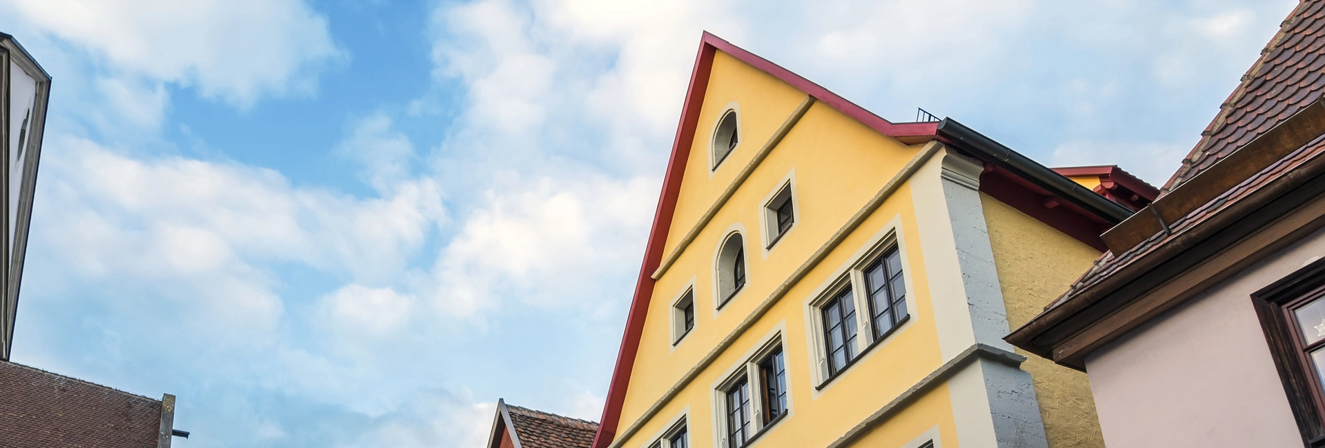 Gelbes Haus mit rotem Dach und mehreren Fenstern vor einem teilweise bewölkten Himmel neben anderen Gebäuden mit Ziegeldächern.