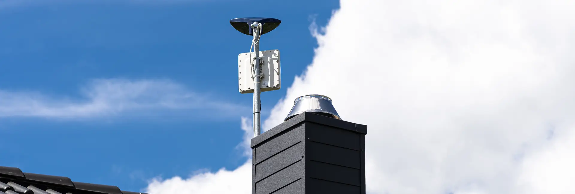 Auf einem Dach sind eine kleine Satellitenschüssel und eine Entlüftung montiert, im Hintergrund sind ein blauer Himmel und weiße Wolken zu sehen.