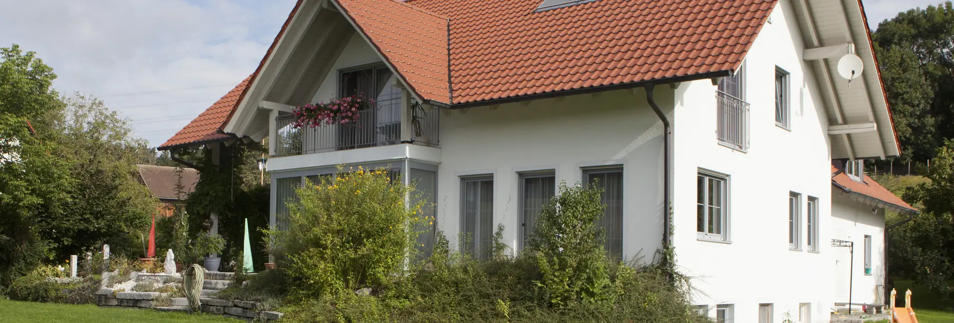 Ein weißes Haus mit rotem Satteldach, großen Fenstern und einem Garten mit Sträuchern und Blumen im Vordergrund.