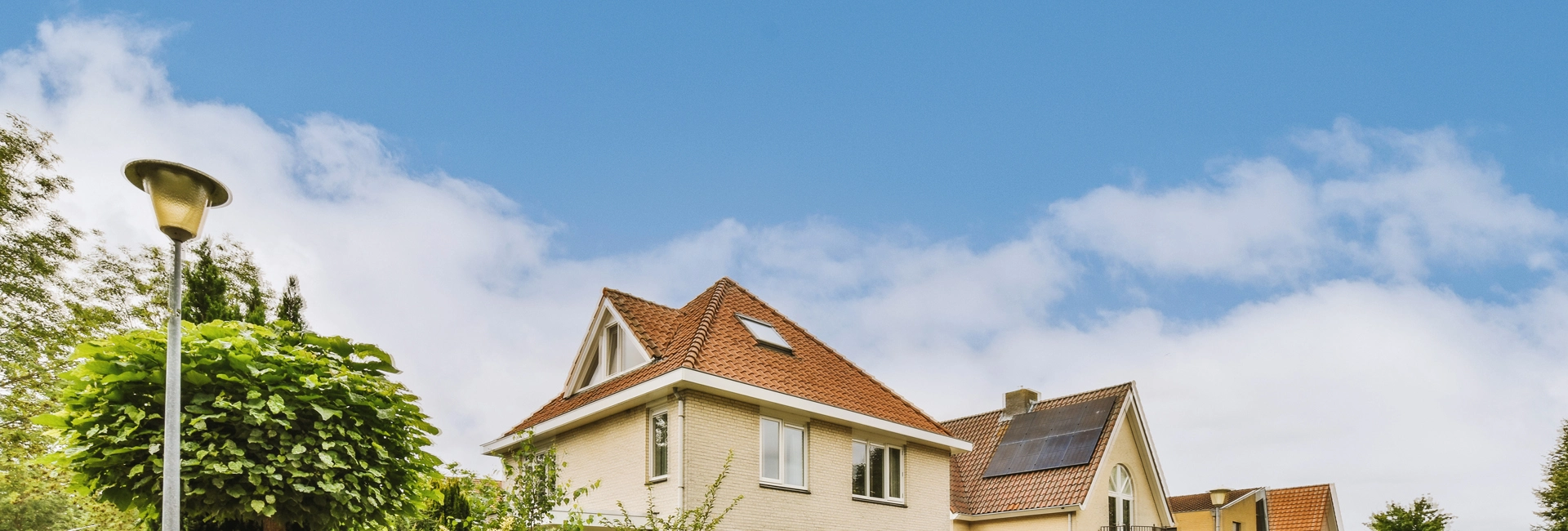 Unter einem blauen Himmel mit vereinzelten Wolken ist ein beiges Haus mit rotem Ziegeldach und Solarmodulen zu sehen, umgeben von Grün und einer Straßenlaterne.